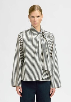 Kvinde med blonde hår iført sort-hvidt diagonalt stribet bluse med halsbånd og sorte bukser mod hvid baggrund.