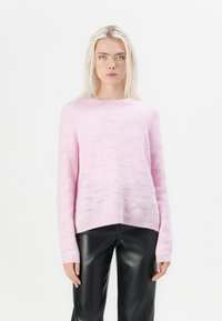 Model in een lichtroze, getextureerde trui en zwarte leren broeken, staand tegen een effen witte achtergrond, met een neutrale uitdrukking.