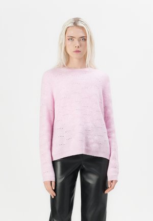 Model in een lichtroze, getextureerde trui en zwarte leren broeken, staand tegen een effen witte achtergrond, met een neutrale uitdrukking.