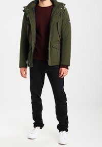 Oliivinvihreä puffer-takki, jossa on vetoketjullinen etuosa, säädettävä huppu ja useita taskuja, päällä marooninvärinen villapaita ja mustat housut.