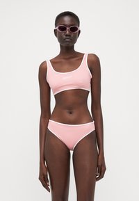 Rozā bikini komplekts ar baltu apmalīti, kurā ir izgriezts augšdaļa un zemas jostras apakšdaļas. Modelis valkā lavandas krāsas saulesbrilles pret neitrālu fona.