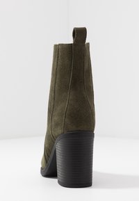 Stivale alla caviglia in suede verde oliva con tacco a blocco nero impilato e linguetta sul retro, mostrato da un angolo posteriore su sfondo bianco.
