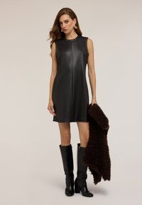 Vestido de cuero negro sin mangas con tachuelas plateadas en el frente y el escote, combinado con un chal texturizado marrón y botas negras hasta la rodilla.