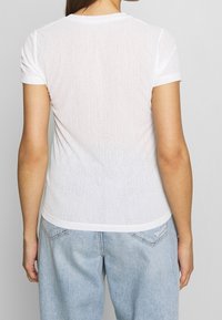 White, short-sleeve T-shirt made of thin, slightly sheer fabric, featuring a round neckline and a fitted shape. Worn with light blue jeans.