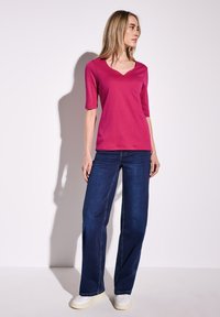 Fuksjowa bluzka z dekoltem w serek i rękawami do łokcia, zestawiona z szerokimi ciemnoniebieskimi dżinsami oraz białymi trampkami. Gładka faktura i luźny krój.