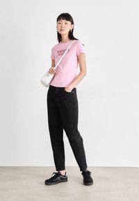 Rozā īsām piedurknēm T-krekls ar uzrakstu "tommy" sarkanā krāsā, kombinēts ar melnām taisnām biksēm un melniem sporta apaviem, tur rokās baltu mācību somu.