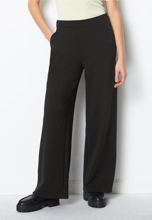 Frau trägt schwarze Hosen mit weitem Bein und schwarze klobige Loafers, steht mit einer Hand in der Tasche vor einer weißen Wand.