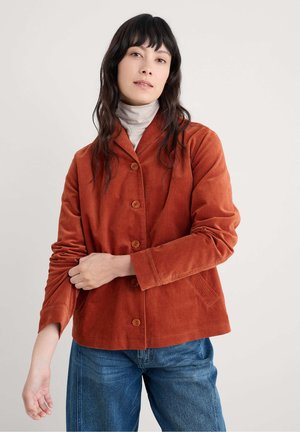 Brændt orange kordfløjl jakke med krave, fem knapper og to forlommer. Lange ærmer og afslappet pasform, parret med blå jeans.