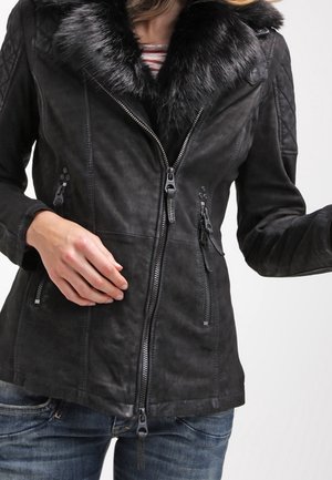 Femme portant une veste en daim noire avec un grand col en fourrure noire, fermée par une fermeture éclair à l'avant, associée à un jean bleu et un haut rayé en dessous.