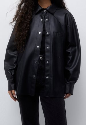 Person wearing a black faux leather button-up shirt with long sleeves over a black top and high-waisted black pants against a gray background.