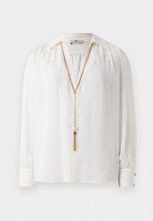 Valkoinen pitkähiha blouse, jossa on hurstutetut hartiat, terävä kaulus, napitettu hihansuu ja pitkä kultaketju, jossa on tupsuyksityiskohta.