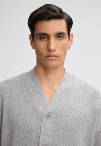 Grauer Strickcardigan mit einem V-Ausschnitt, ausgestattet mit zwei lila Knöpfen. Der Stoff wirkt weich und strukturiert.