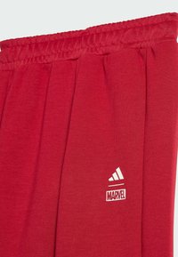 Röda sweatpants i mjukt tyg med ribbad midja och vita tryckta logotyper för Adidas och Marvel vid underbenet.