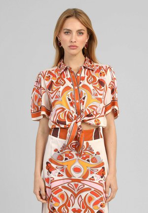 Femme portant un chemisier à manches courtes noué assorti avec jupe à motifs orange, marron et rose, debout devant un fond gris uni.