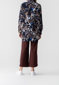 Kwiatowa koszula z długim rękawem w granatowym kolorze z białymi, brązowymi i niebieskimi wzorami, zestawiona z brązowymi spodniami typu wide-leg i białymi trampkami.