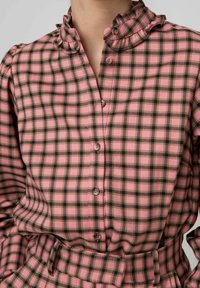 Camicia a quadri rosa e nera con colletto arricciato, bottoni sul davanti e vita stringata, caratterizzata da una morbida texture di tessuto.