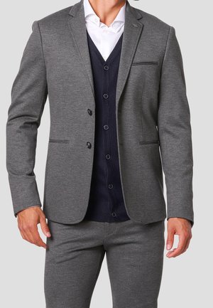 Un homme portant un costume gris avec un blazer à deux boutons, un cardigan bleu marine et une chemise blanche, se tenant devant un fond uni.