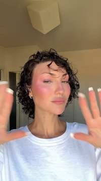 Lockar med tydlig textur; bär en vit skjorta; ljus rosa rouge på kinderna; lyfta händer nära ansiktet.