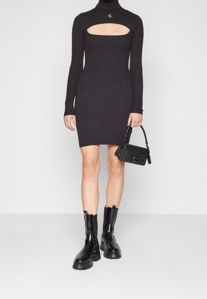 Czarna dzianinowa sukienka z długimi rękawami i kwadratowym wycięciem, w zestawieniu z czarnymi botkami za kostkę oraz małą czarną torebką.