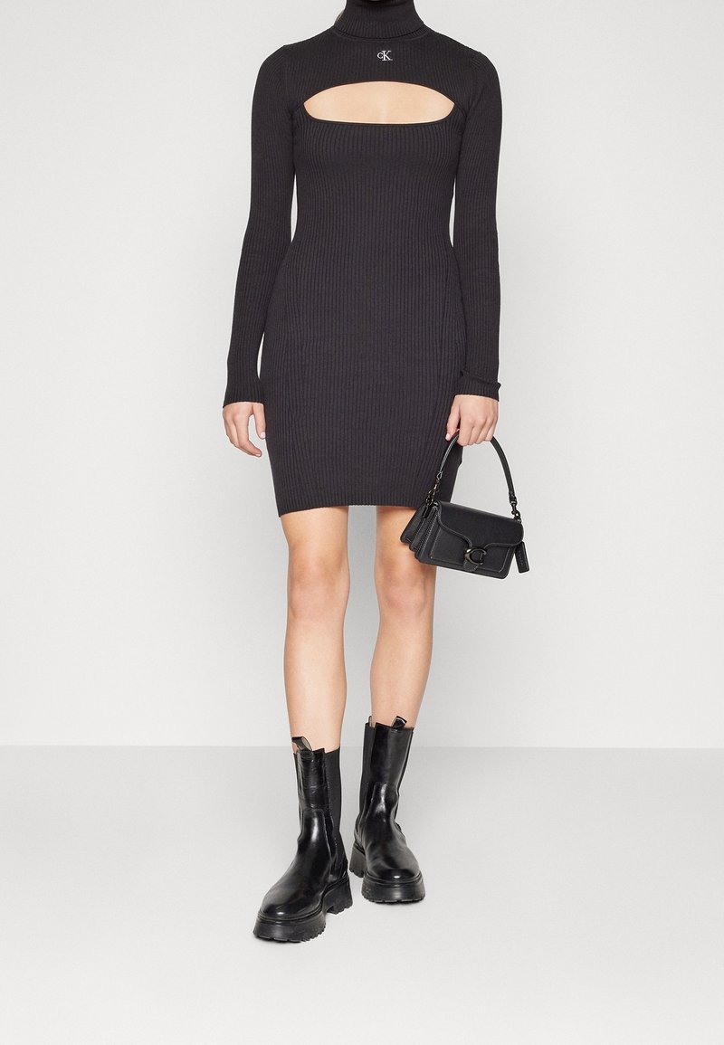 Robe pull noire côtelée avec une découpe carrée, manches longues, associée à des bottines noires et un petit sac à main noir.