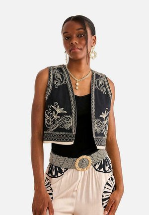 Mujer con chaleco negro bordado sobre camiseta negra, cinturón con hebilla tejida y pantalones crema con estampados geométricos negros.