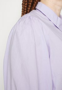 Helle lila Bluse mit einem subtilen Glanz, strukturiertem Kragen und gefältelten Puffärmeln. Glatter Stoff mit minimalen Nähdetails.