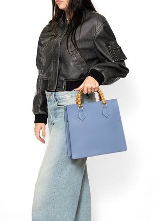 Vrouw draagt een zwart leren jas en lichtblauwe spijkerbroek, houdt een blauwe handtas met handvatten in bamboestijl naast zich vast.