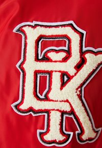Rood stof met een gestructureerd logo in een wit, vachtachtig materiaal, omrand met zwarte en rode stiksels, met de letters "P" en "K."
