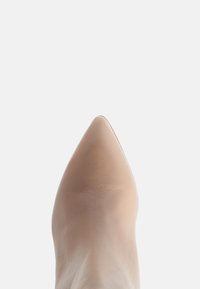 Vârful unei pantofi din piele bej ascuțită pe un fundal gri deschis, punând în evidență textura și forma.