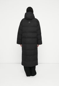 Sort polstret frakke med et langt, oversized design, stor hætte og vandret kviltede paneler. Har et logo på ryggen. Glat tekstur.