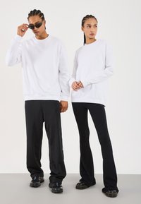 Two models wearing white sweatshirts; one in black wide pants and sunglasses, the other in black flared ribbed pants and boots.