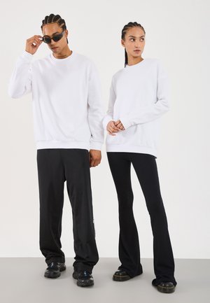 Deux modèles portant des sweat-shirts blancs ; l'un en pantalon large noir et lunettes de soleil, l'autre en pantalon évasé noir à côtes et bottes.