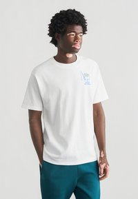 Λευκό βαμβακερό μπλουζάκι με κοντά μανίκια, με μπλε γραφικό σχέδιο και κείμενο στην αριστερή πλευρά του στήθους. Συνδυασμένο με παντελόνια σε τιρκουάζ αποχρώσεις.