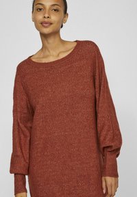 Maglione di colore ruggine in knit con scollatura ampia, maniche ampie e polsini a coste, che mostra un tessuto testurizzato e una vestibilità rilassata.