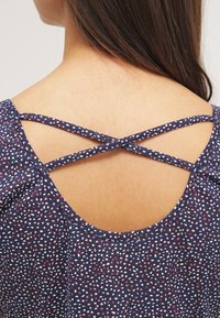 Vista trasera de una mujer que lleva un vestido azul marino con motas blancas y rojas y tirantes finos cruzados.