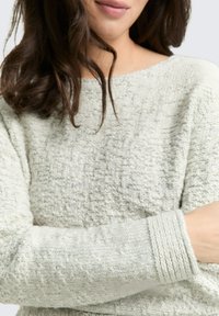 Pull en gris clair avec un tissu tricoté texturé. Présente un col large et des poignets côtelés. Apparence douce et confortable avec des motifs subtils.