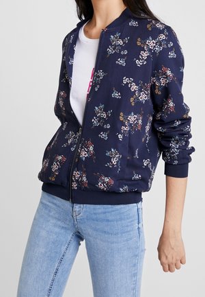 Vrouw draagt een marineblauwe bloemige zip-up jas over een wit T-shirt, gecombineerd met lichtblauwe spijkerbroek, staand met één arm ontspannen.