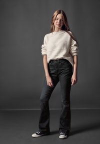 Suéter oversized de color beige claro con textura y mangas enrolladas, combinado con jeans acampanados negros de cintura alta y zapatillas negras. Fondo simple.