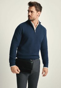 Granatowy sweter z zamkiem na kołnierzu, w zestawie z szarymi spodniami. Model trzyma czarną skórzaną teczkę. Widoczny subtelny wzór tekstury.