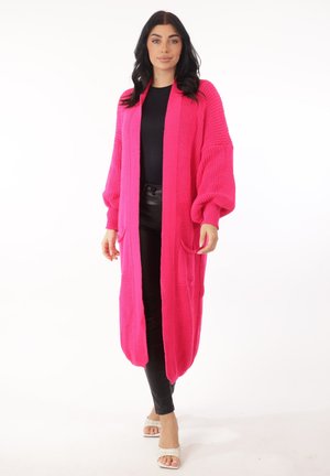 Mujer vistiendo un cárdigan largo de punto de color rosa brillante con mangas abullonadas sobre un top y pantalones negros, posando contra un fondo blanco.