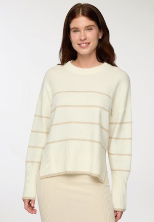 Donna con lunghi capelli castani, che indossa un dolcevita color crema con sottili strisce beige e una gonna a coste coordinata, sorridente su uno sfondo bianco.