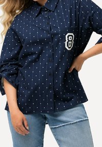 Navy blue button-up shirt featuring white polka dots, a chest pocket with a large number "8" in white, and rolled-up sleeves.