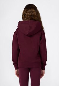 Enfant avec de longs cheveux ondulés portant un sweat à capuche maroon uni et un pantalon assorti, se tenant de dos devant un fond blanc.