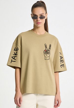 Donna che indossa una t-shirt beige oversize con grafica del simbolo della pace e parole "Faith," "Love," "Peace," "Joy," "Hope," e "Take Care" sulle maniche.