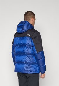 Niebieska kurtka puchowa z czarną górną częścią, z kapturem, pikowanym wzorem i elastycznymi mankietami. Logo na lewym ramieniu.