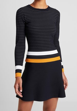 Femme portant un haut noir moulant à manches longues avec des rayures blanches et orange et une jupe noire sur un fond uni.