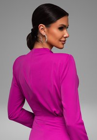 Fuchsia langjurk met hoge hals en een aansluitende taille. Glad stoffen textuur en een ritsluiting aan de achterkant. Zilveren ringoorbellen.