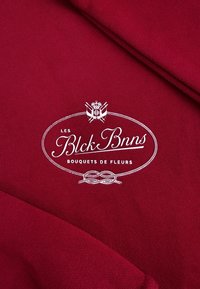 Bordeaux textiel met een wit geborduurd logo binnen een ovale lijst, versierd met decoratieve touwaccenten en bloemenmotieven.