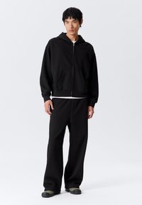 Sudadera negra con cremallera, bolsillo frontal y corte holgado, combinada con pantalones de chándal negros. El modelo lleva calzado negro y verde.