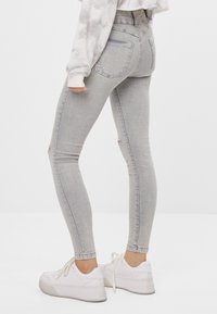 Jeans ajustados grises con detalles desgastados en las rodillas, cintura alta y costura en contraste. Combinados con zapatillas blancas para un look casual.